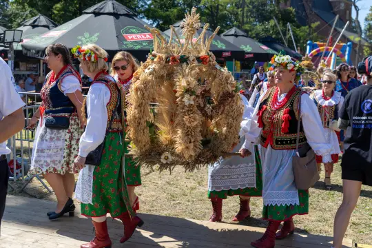 Kobiety w tradycyjnych strojach ludowych niosą wiechę z kwiatów i zboża podczas festiwalu.