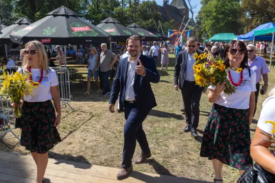 Mężczyzna w garniturze idzie wśród kobiet z kwiatami, w tle festyn z namiotami.
