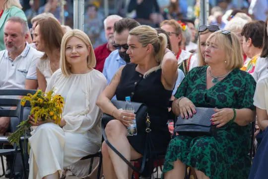 Kobiety w eleganckich strojach siedzą na widowni, jedna trzyma bukiet kwiatów.