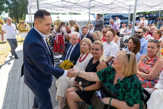 Mężczyzna w garniturze wita się z kobietą w zielonej sukience, w tle zgromadzenie ludzi.