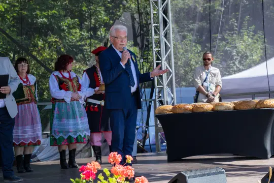 Mężczyzna w garniturze przemawia na scenie, obok niego stoją osoby w tradycyjnych strojach ludowych.
