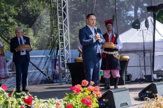 Mężczyzna w garniturze przemawia na scenie, obok tradycyjnie ubrani uczestnicy z chlebem.