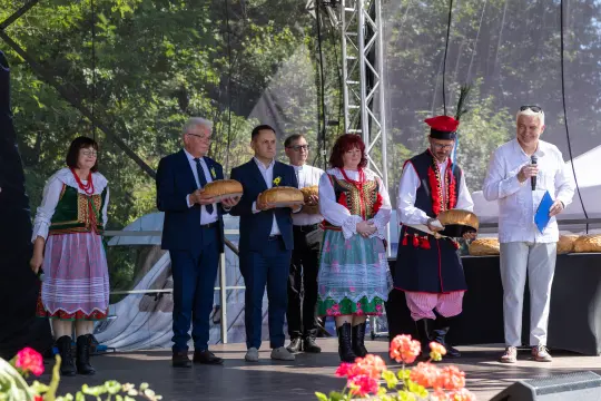 Grupa osób w tradycyjnych strojach ludowych trzyma chleb na scenie podczas wydarzenia.