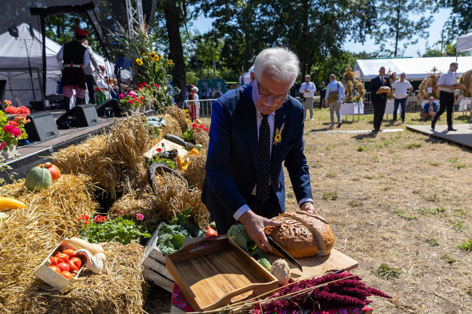 Mężczyzna kroi chleb na tle dekoracji z warzywami i słomy podczas festynu.  Autor: Bogusław Świerzowski