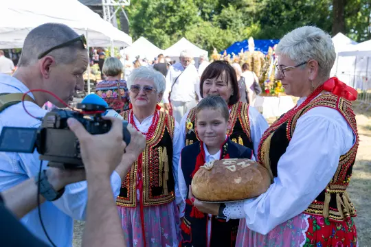 Grupa osób w tradycyjnych strojach ludowych trzyma chleb na festynie.