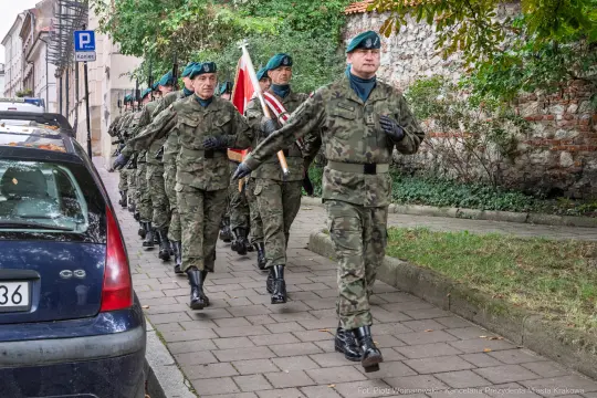 Żołnierze w mundurach wojskowych maszerują ulicą, niosąc flagę.