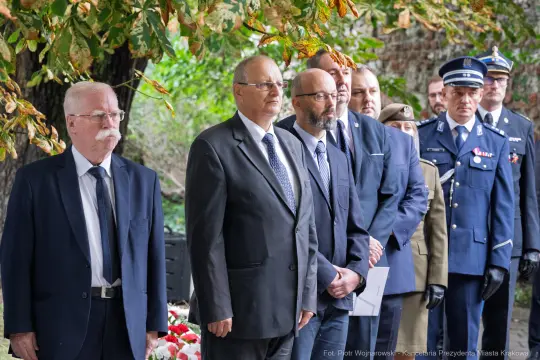 Uroczystość złożenia kwiatów przez grupę mężczyzn w garniturach i mundurach.