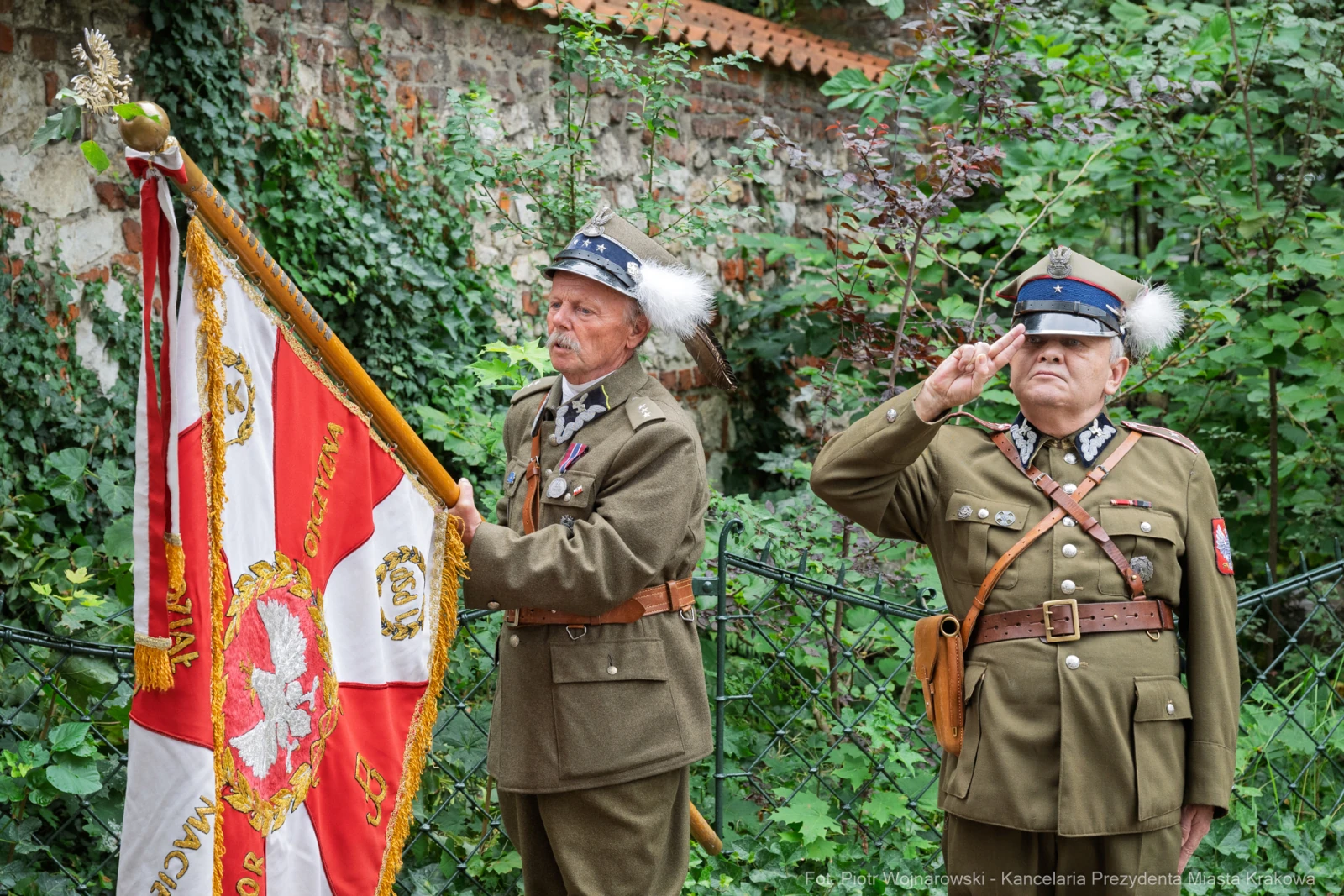 Dwaj mężczyźni w mundurach wojskowych z flagą, jeden salutuje. Tło z zielenią i historycznymi budynkami.  Autor: Piotr Wojnarowski