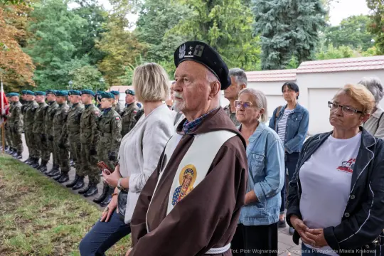 Uczestnicy ceremonii, w tym weteran, stoją wśród żołnierzy i innych osób.