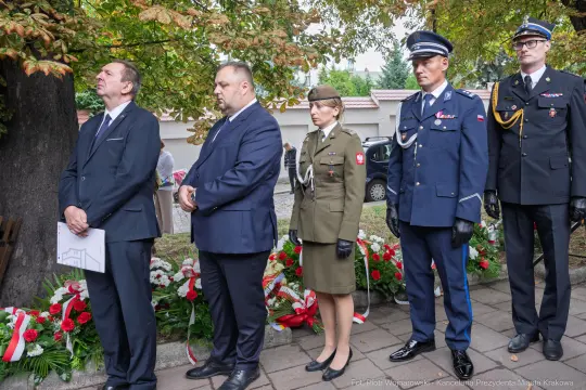 Uczestnicy ceremonii złożenia kwiatów, w mundurach i garniturach, przy pomniku.