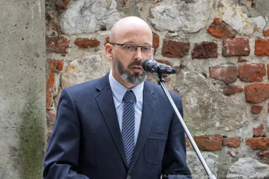 Mężczyzna w okularach przemawia do mikrofonu, w tle ceglana ściana.