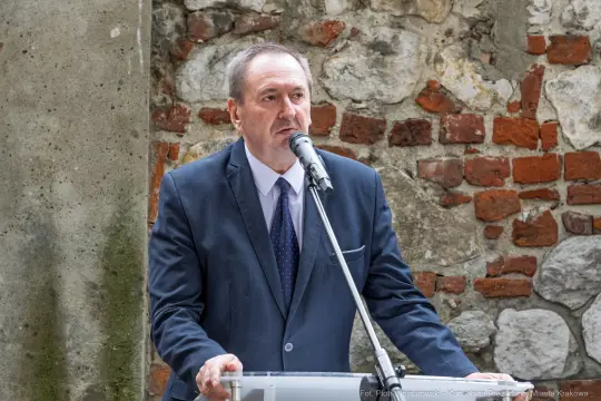 Mężczyzna w garniturze przemawia do mikrofonu, w tle ceglana ściana.