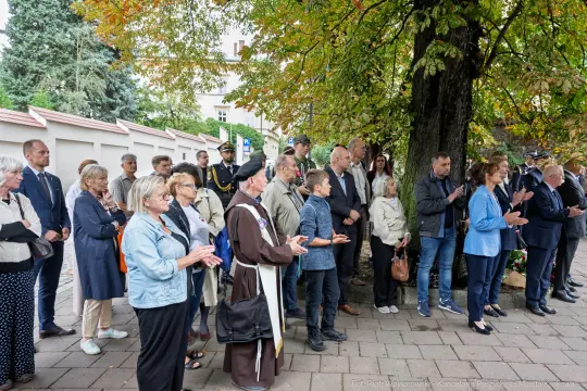 Grupa ludzi w różnym wieku, w tym duchowny, zgromadzona pod drzewem.