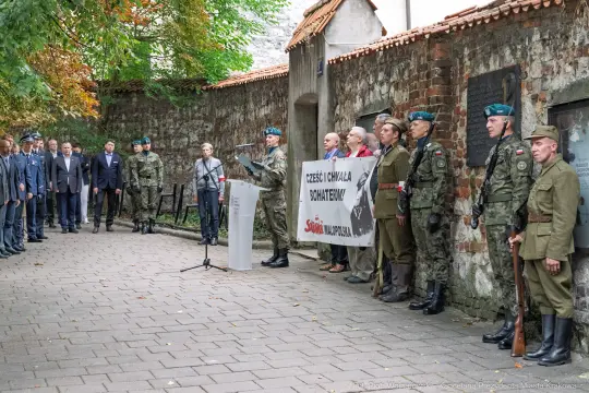 Ceremonia z udziałem żołnierzy i oficjeli, z transparentem "Cześć i chwała bohaterom".