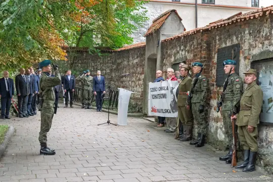 Uroczystość z udziałem żołnierzy i oficjeli, z transparentem w tle.