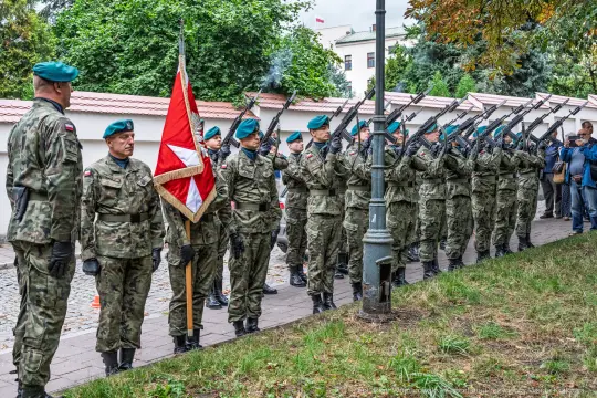 Żołnierze w mundurach wojskowych z bronią w ręku, salutujący z flagą.
