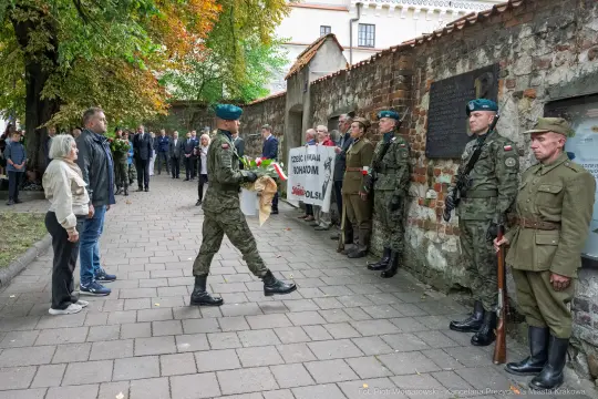 Uroczystość złożenia kwiatów przez żołnierzy w towarzystwie uczestników na tle historycznego muru.