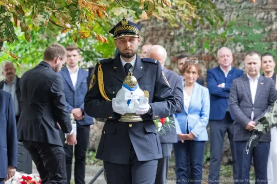 Mężczyzna w mundurze trzyma znicz, wokół zgromadzenie ludzi w tle.
