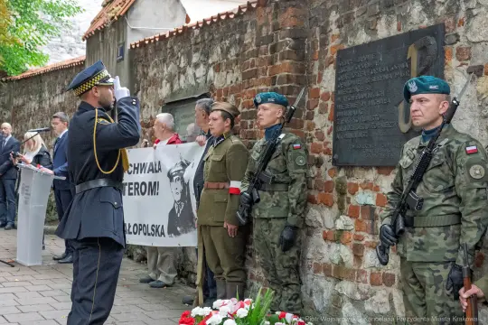 Ceremonia upamiętniająca z udziałem żołnierzy i flagi, z tablicą pamiątkową w tle.