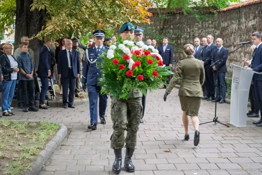Żołnierz niesie wieniec na uroczystości, w tle zgromadzeni goście i przedstawiciele służb mundurowych.