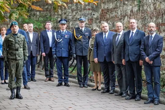 Grupa ludzi w formalnych strojach oraz mundurach, stojących wzdłuż muru.