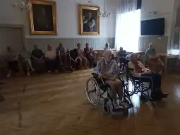 Grupa starszych osób w wózkach inwalidzkich i na krzesłach w jasnym pomieszczeniu.