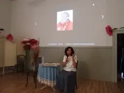 Kobieta siedząca przy stole, z tłem w postaci zdjęcia starszej kobiety.