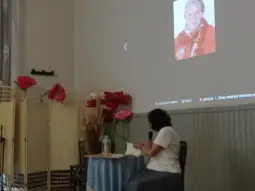 Kobieta siedząca przy stole, obok kwiaty i ekran z portretem starszej kobiety.