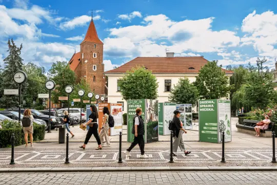 Ulica z ludźmi, zielenią i plakatami informacyjnymi, w tle wieża kościoła.