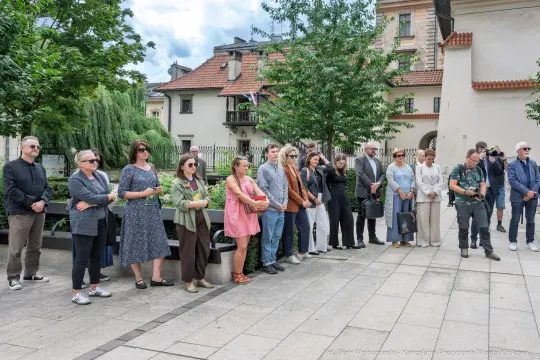 Grupa ludzi stojąca w parku, niektórzy trzymają kwiaty, w tle budynki i zieleń.