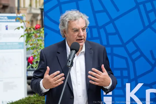 Mężczyzna w okularach, przemawiający do mikrofonu, z tłem w kolorze niebieskim.