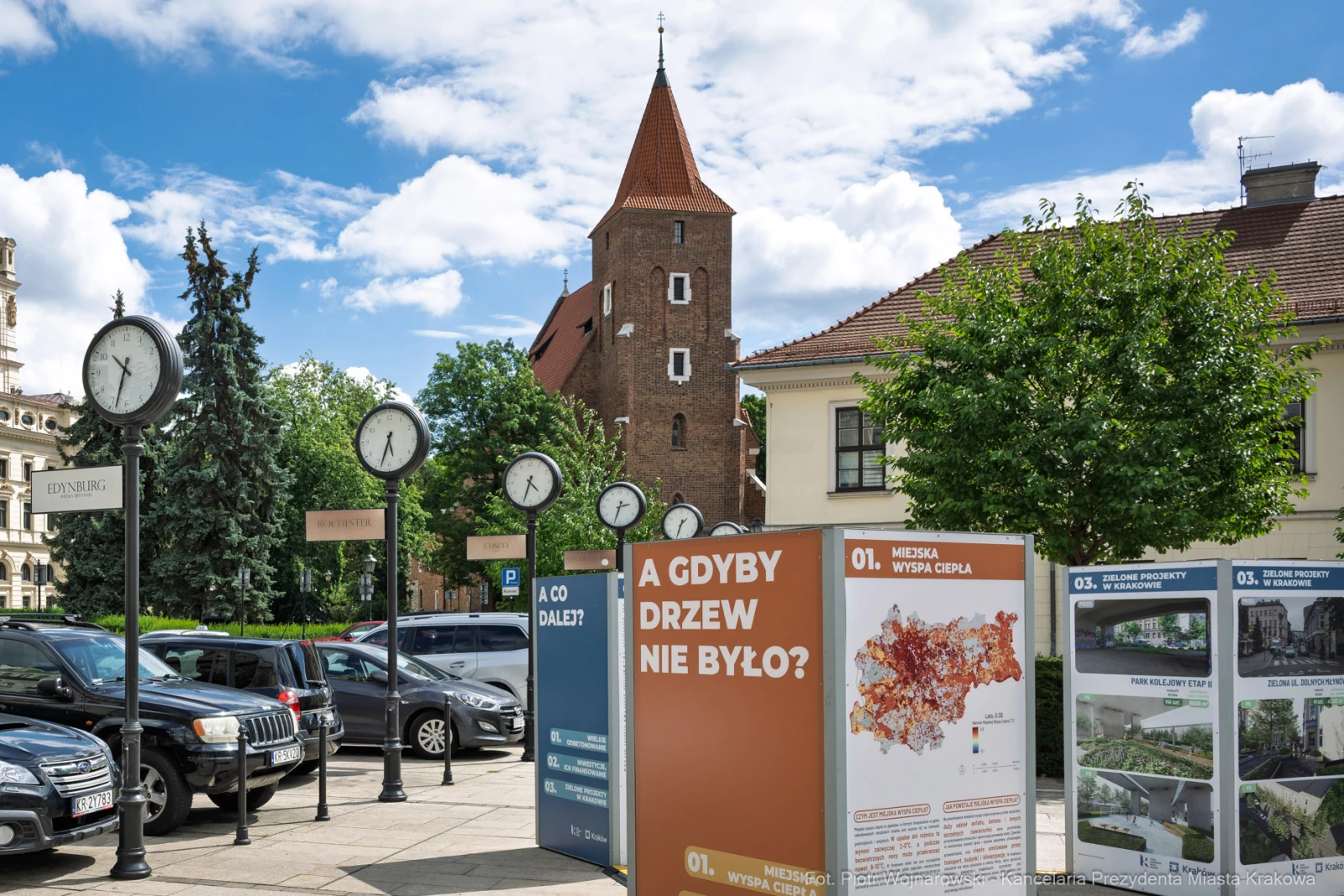 Zegarowe słupy i tablice informacyjne w miejskim krajobrazie z wieżą w tle.  Autor: Piotr Wojnarowski