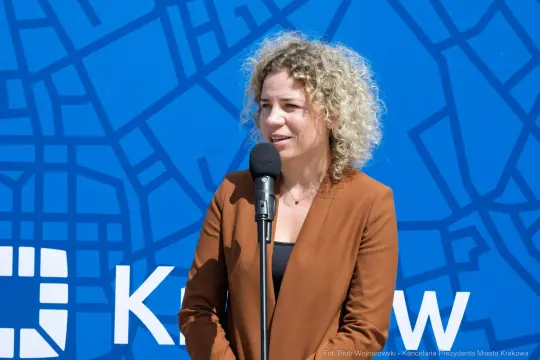 Kobieta z kręconymi włosami przemawia do mikrofonu na tle niebieskiego tła z mapą.