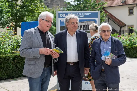 Trzech mężczyzn trzymających ulotki, stojących w miejskim otoczeniu.