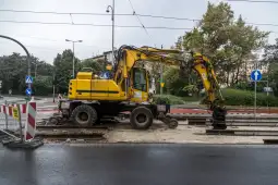 Żółty koparka pracująca przy torach tramwajowych w deszczowej scenerii.