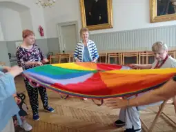 Grupa osób bawi się kolorowym parasolem w sali.