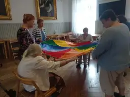 Grupa osób bawi się kolorowym chustą w sali.