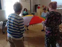 Grupa osób bawi się kolorowym parasolem w sali.