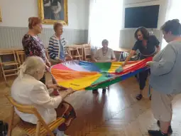 Grupa osób starszych bawi się kolorowym parasolem w sali.