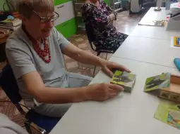Starsza kobieta z okularami trzyma w rękach kolorowe pudełko, uśmiechając się. W tle inne osoby przy stole.