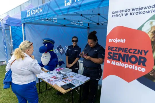 Stoisko informacyjne z policjantkami, maskotką i materiałami dla seniorów w Krakowie.