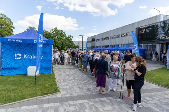 Ludzie w kolejce przed namiotami z logo Krakowa, w słoneczny dzień.