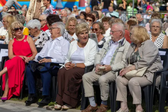 Grupa osób siedzących na krzesłach, wśród nich starsze panie i panowie, w różnorodnych strojach.