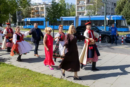 Grupa osób w tradycyjnych strojach ludowych spaceruje po chodniku.