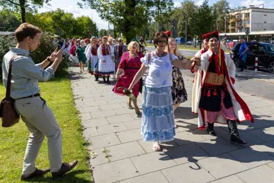 Grupa ludzi w tradycyjnych strojach idzie ulicą, jedna osoba robi zdjęcie.