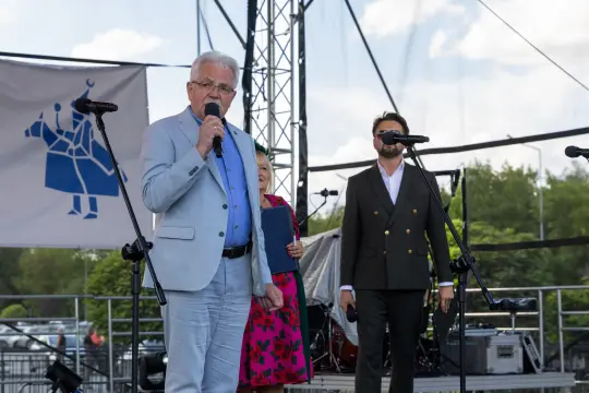 Mężczyzna w jasnym garniturze przemawia do mikrofonu, obok stoi inny mężczyzna i kobieta w kolorowych strojach.