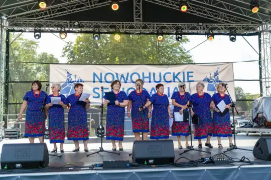 Grupa kobiet w niebieskich sukienkach z czerwonymi akcentami śpiewa na scenie podczas festiwalu.