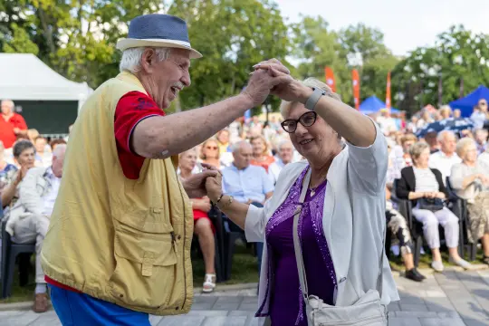 Starszy mężczyzna tańczy z kobietą w okularach, w tle tłum ludzi na wydarzeniu plenerowym.