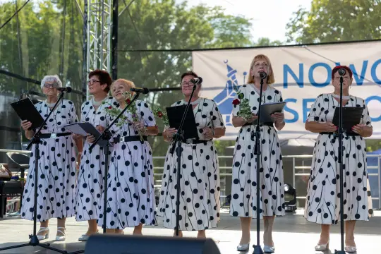 Grupa kobiet w białych sukienkach w czarne kropki śpiewa na scenie podczas wydarzenia.