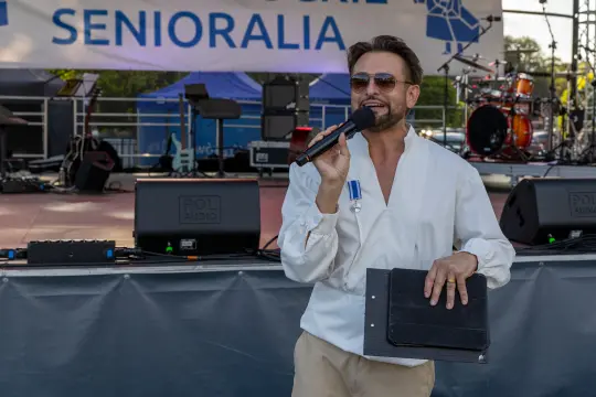 Mężczyzna w białej koszuli trzyma mikrofon i notatnik, prowadzi wydarzenie na scenie.